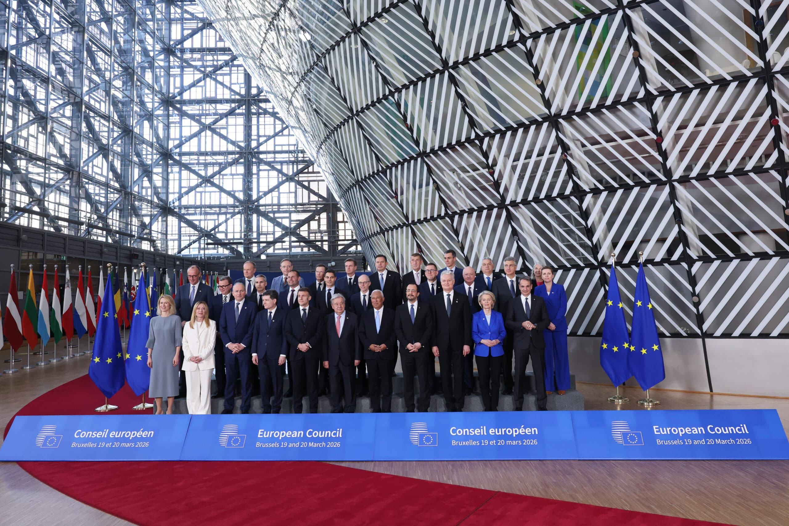 A large group of formal-looking individuals stand together for a photo during the European Council meeting in Brussels on March 19 and 20, 2026. They are positioned in front of a modern, geometric architectural backdrop with EU flags and various national flags lined up on the left side.