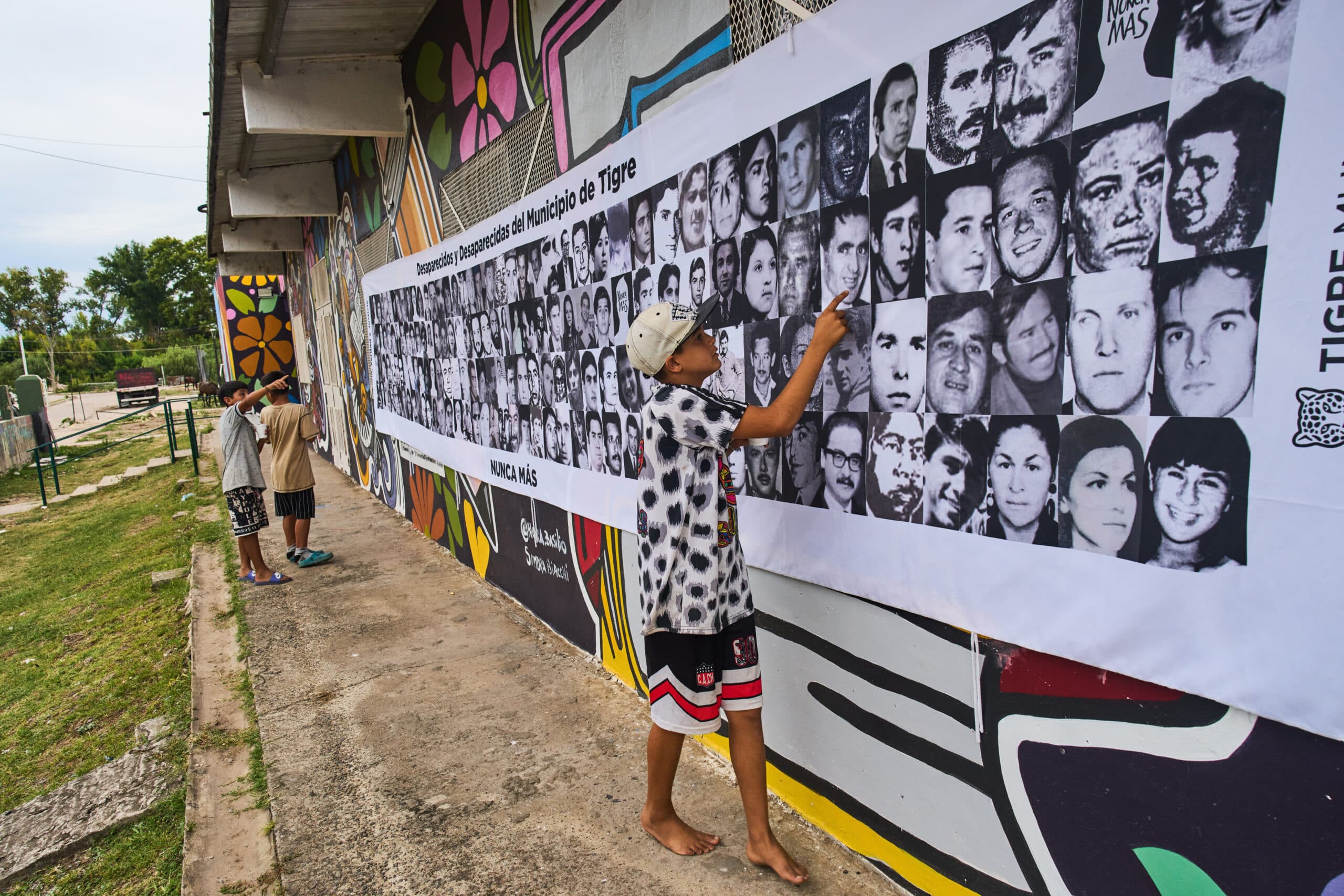 A young person points to a large poster displaying black and white photographs of missing people, titled "Desaparecidos y Desaparecidas del Municipio de Tigre." The poster is attached to a colorful mural on an outdoor wall. Another person is visible in the background observing the display.