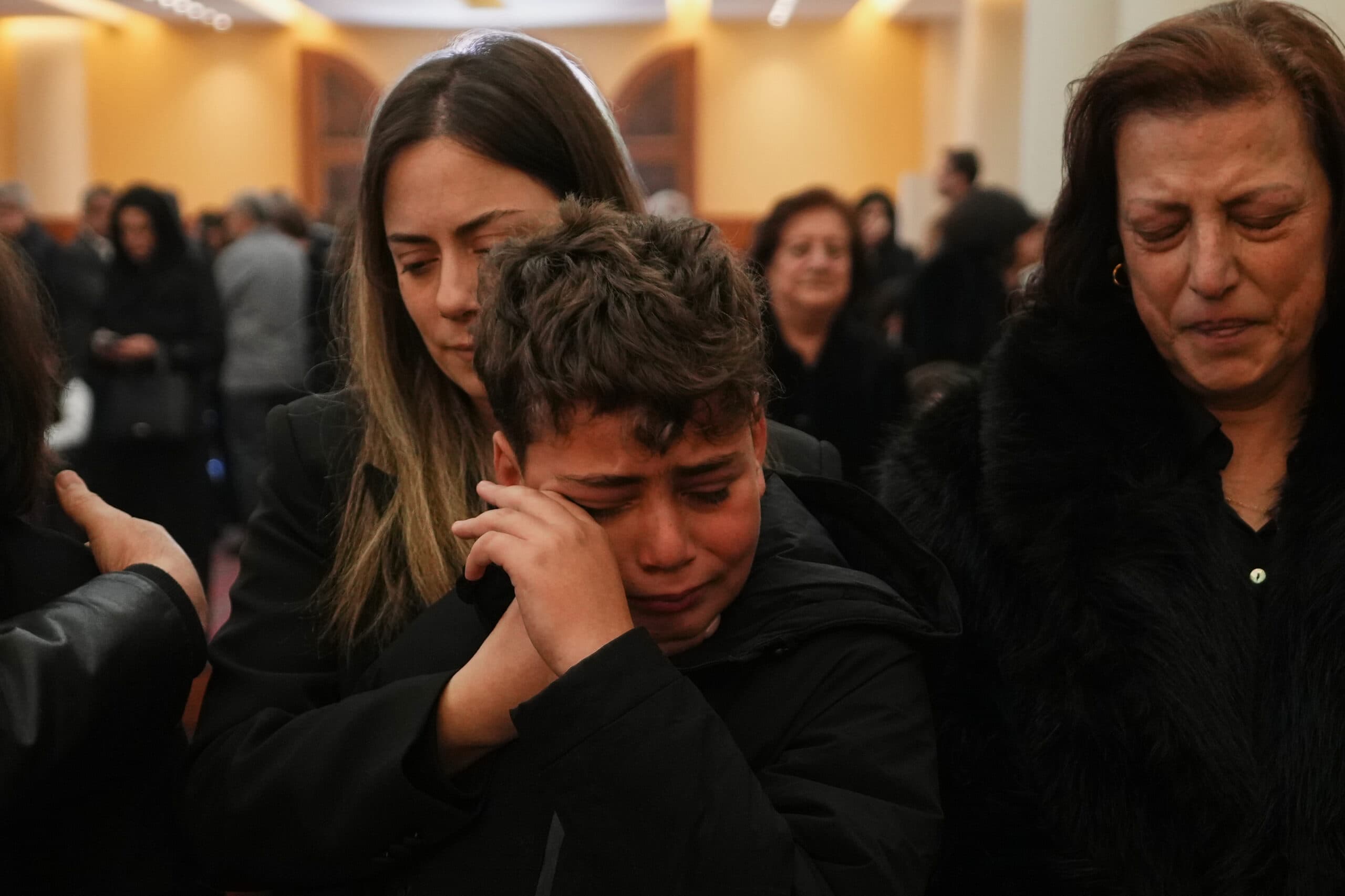 A young boy crying and being comforted by a woman at a solemn indoor gathering, surrounded by other people appearing sad or thoughtful in the background.