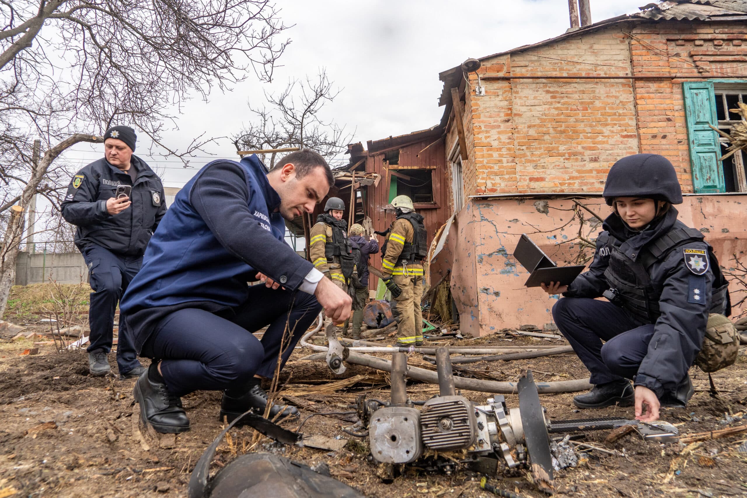 Police and emergency responders inspect debris near a damaged building, likely in a conflict zone.