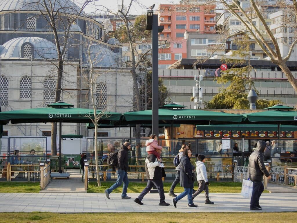 People walking along a path in front of a cafe with green awnings. In the background is a historic building with domes and decorative windows. Leafless trees and modern buildings are also visible.