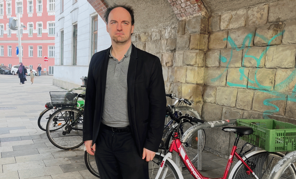 A man in a black jacket and gray polo shirt stands near bicycles parked against a graffiti-covered stone wall. A pink building is visible in the background, along with pedestrians on the street.