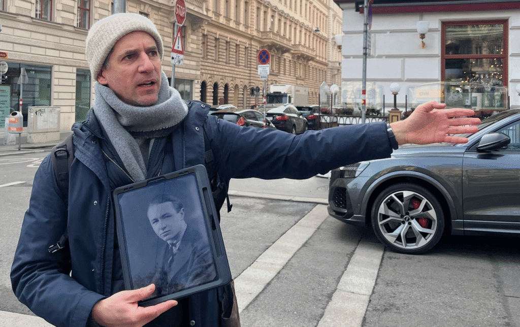 A person wearing warm clothing gestures while holding a tablet displaying a black and white photograph of another person. They stand on a city street with a car and ornate buildings in the background, suggesting a historical tour.