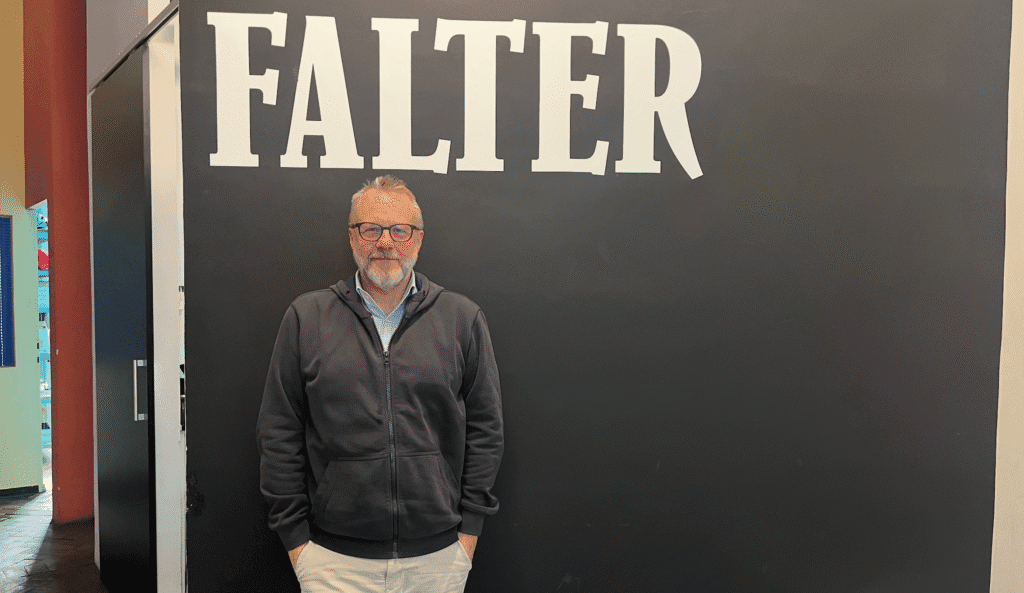 A man wearing glasses and a dark zip-up hoodie stands in front of a black wall with the word "FALTER" written in large white letters.