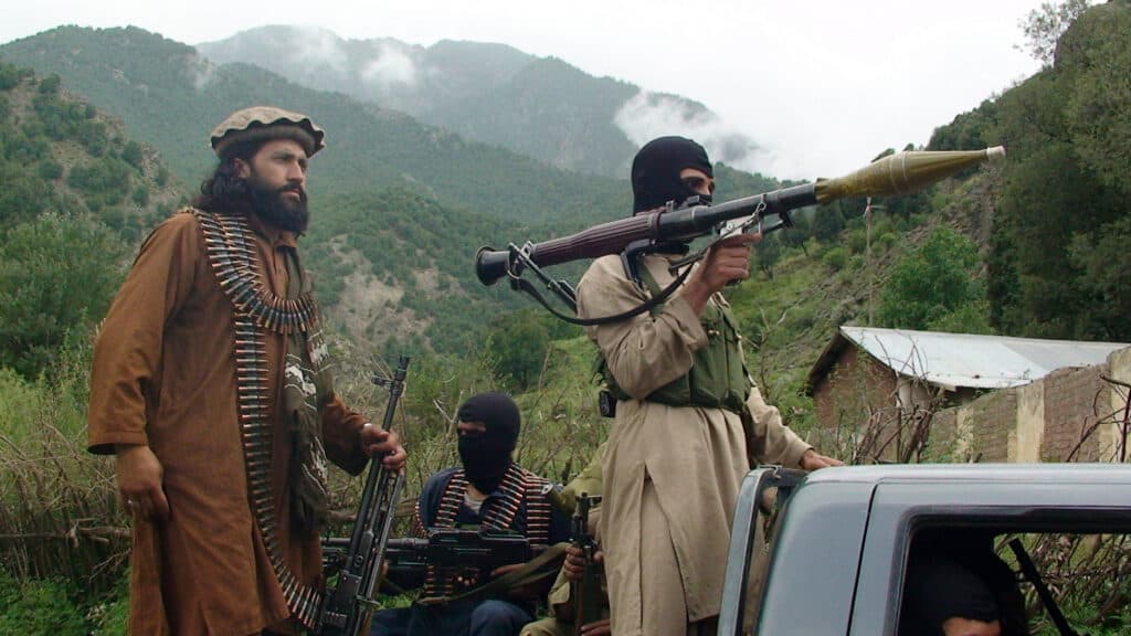Men dressed in military-style clothing, one holding a rocket-propelled grenade launcher, stand outdoors in a mountainous and forested area.