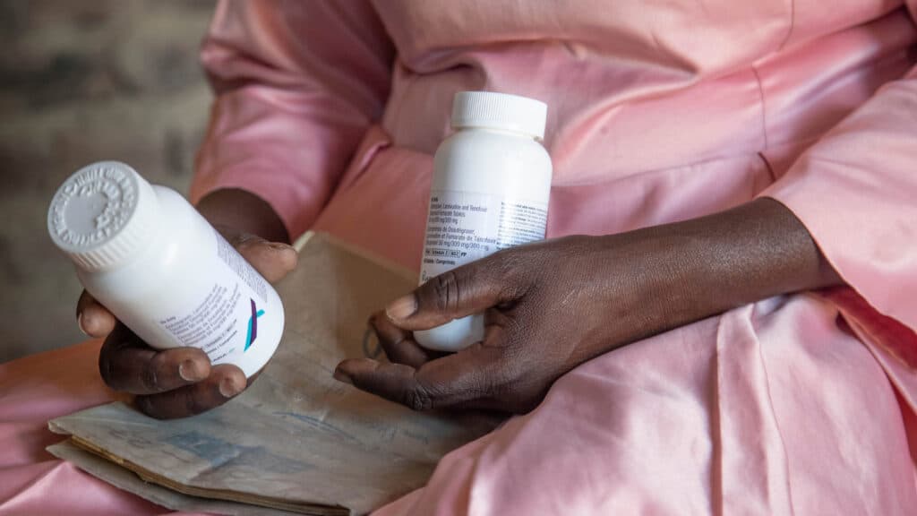 A person in a pink garment holding two white medication bottles labeled with text, one in each hand, while seated.
