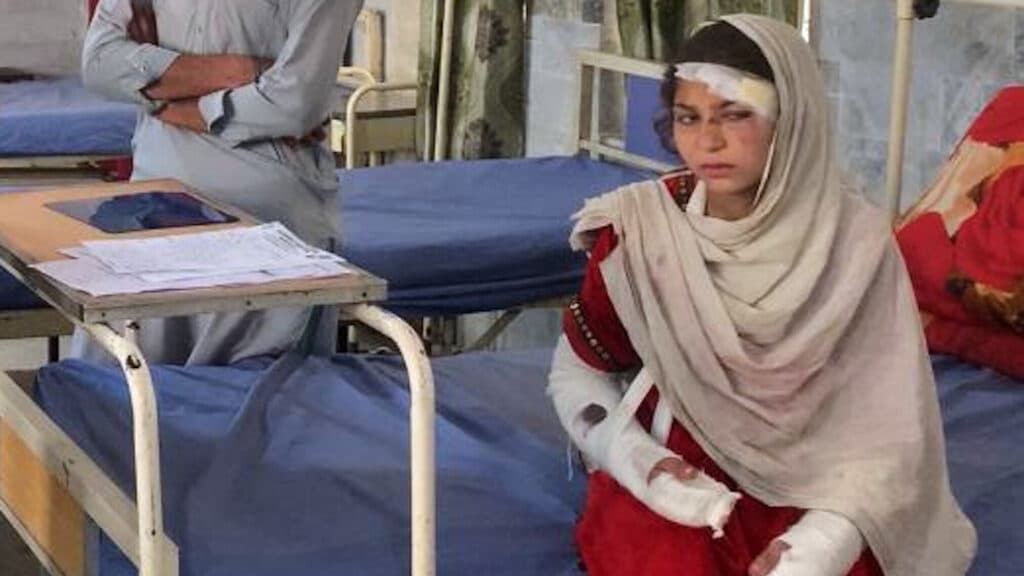 A young girl with bandages on her head and arms sits on a hospital bed, appearing injured. She is wearing a red dress with a beige shawl, and there is a person standing nearby with crossed arms, partially visible.