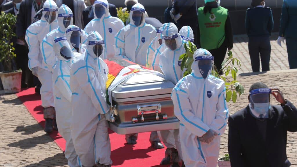 A group of people dressed in white protective suits and face shields carrying a coffin draped with a flag, walking on a red carpet during a formal procession. Other individuals in ceremonial uniforms are visible in the background.