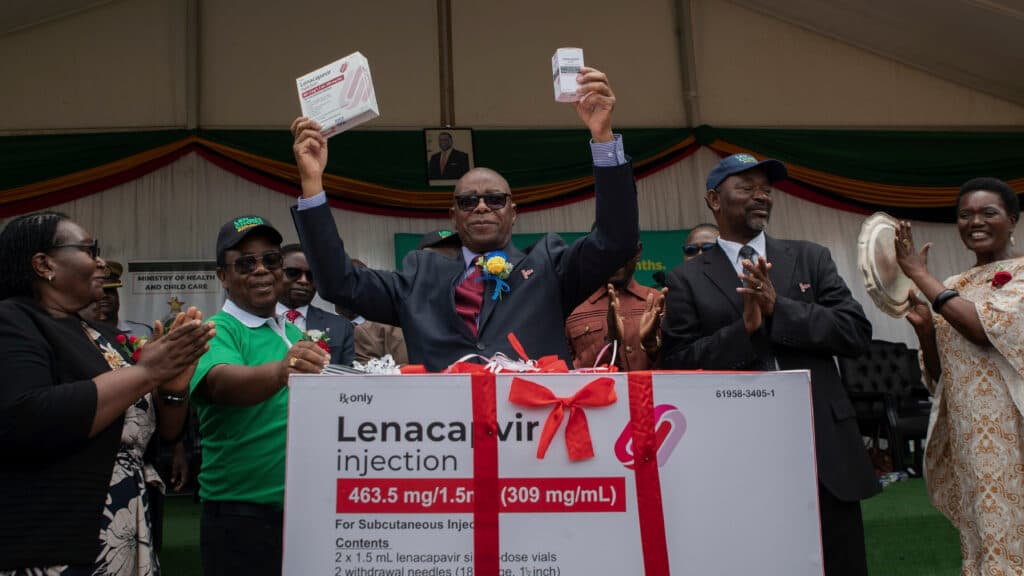 A man in a suit holds two boxes labeled 'Lenacapvir injection' aloft at a public event, surrounded by applauding people in a tent setting with a sign for the Ministry of Health and Child Care in the background.