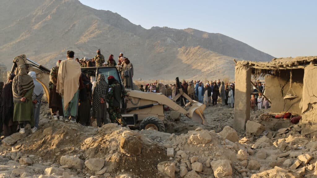 A crowd gathers around rubble and demolition equipment near a damaged dwelling in a mountainous landscape, highlighting a scene of destruction and community presence.