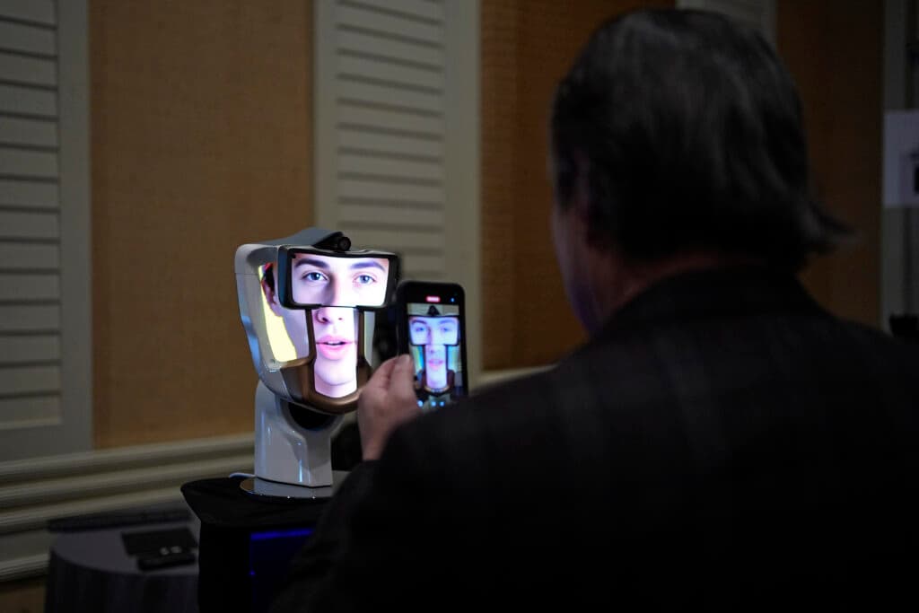 A person takes a picture of a face displayed on a robot-like device using a smartphone in a room with shuttered windows.