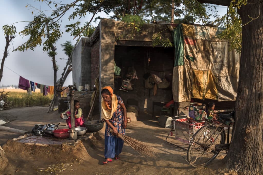 A woman sweeps the dirt in front of a rustic house, with children nearby and household items scattered around.