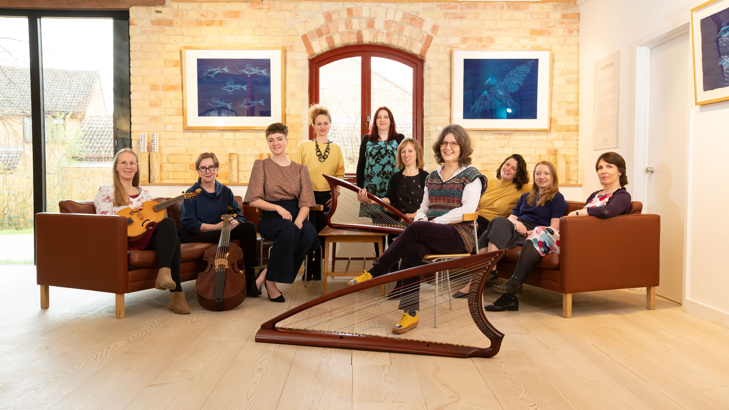 A group of women seated in a bright room, each holding or sitting near musical instruments like a harp and guitar.