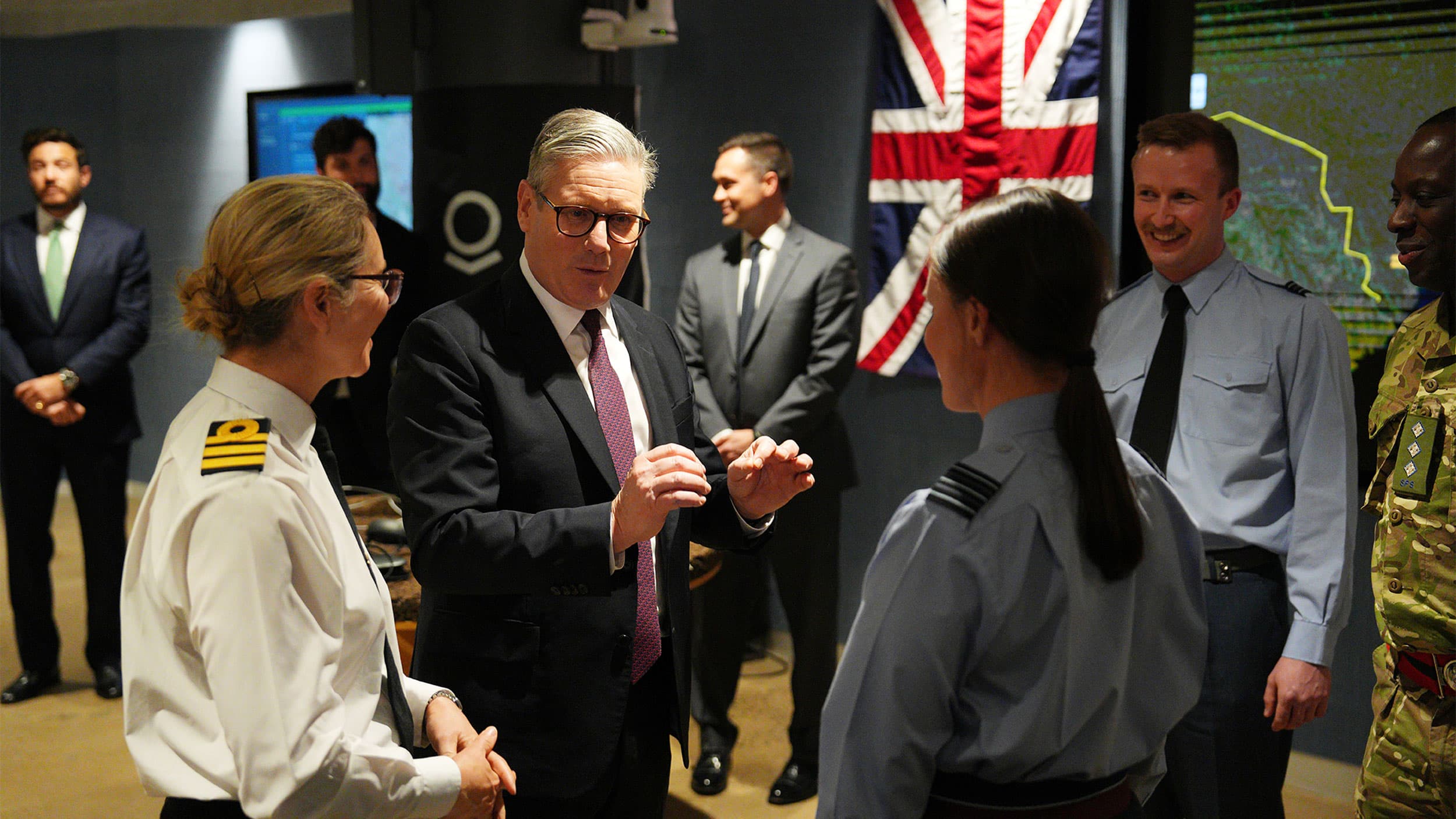 A group of people in military uniforms and formal attire engaged in conversation indoors, with a British flag displayed in the background.