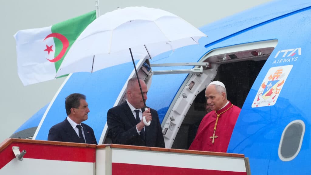 A man in religious attire disembarks a plane under a white umbrella, with the Algerian flag visible in the background.