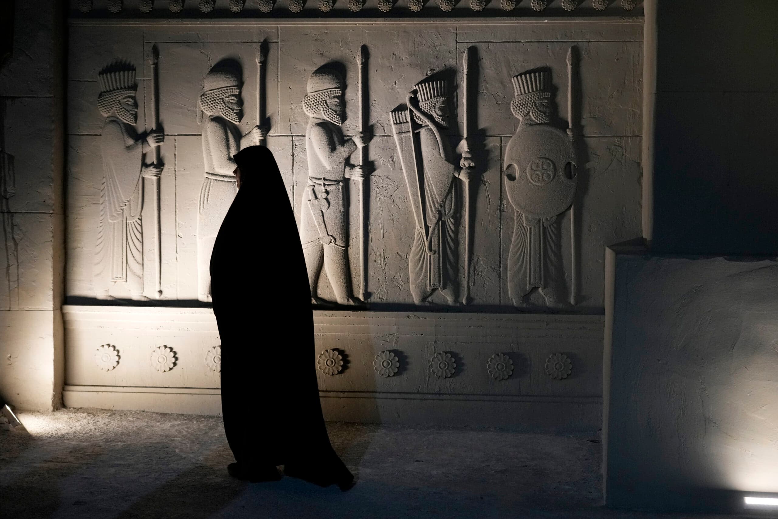 A person in dark clothing walks past a stone wall with ancient warrior carvings, illuminated by soft lighting.