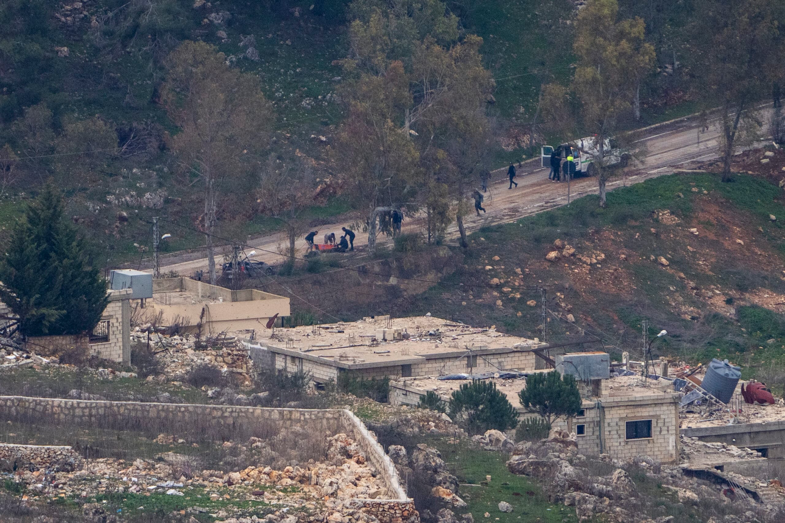 A group of people walking on a dirt path through a rural area with trees and scattered buildings.
