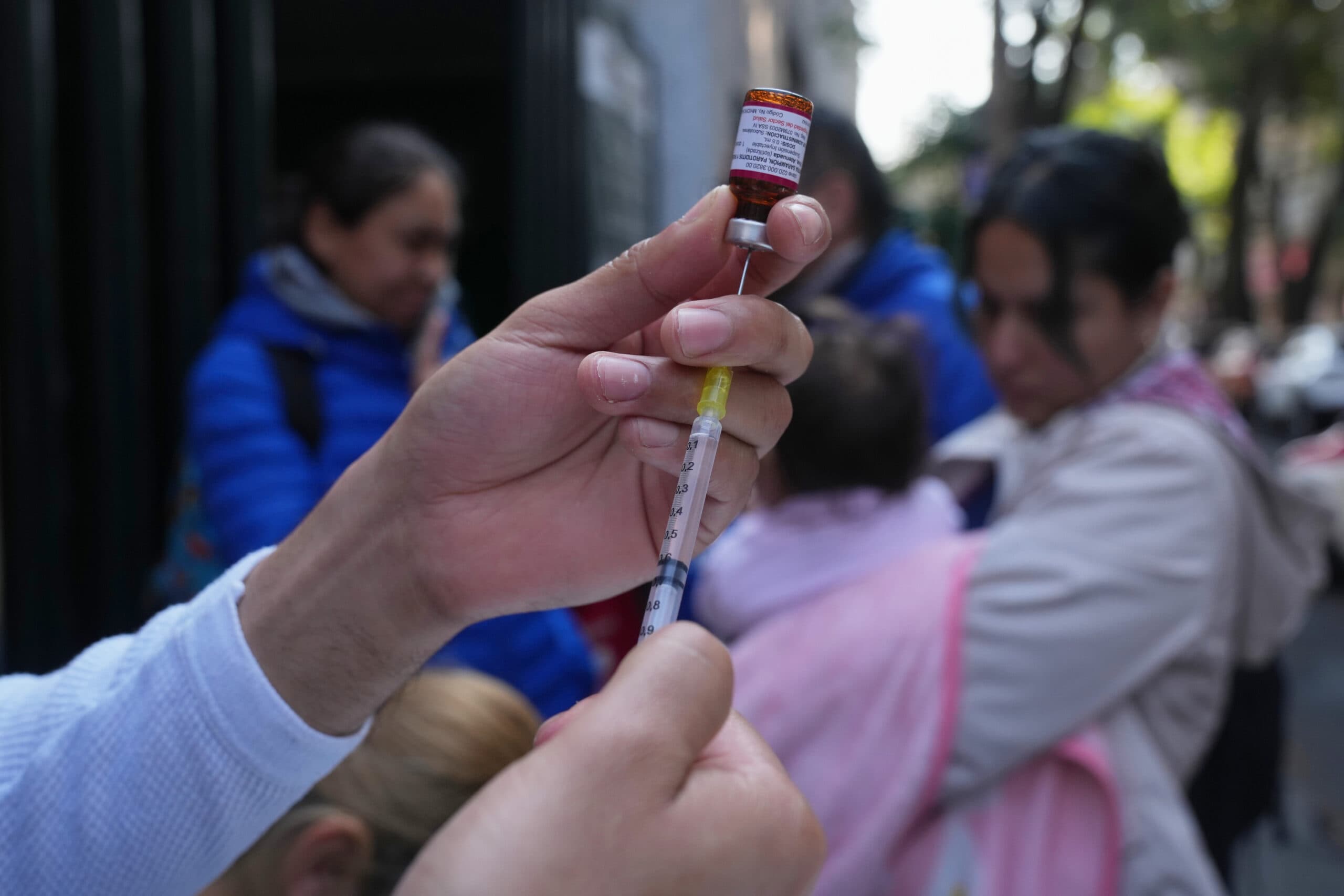 A person fills a syringe with vaccine from a vial, with people and children in the background.