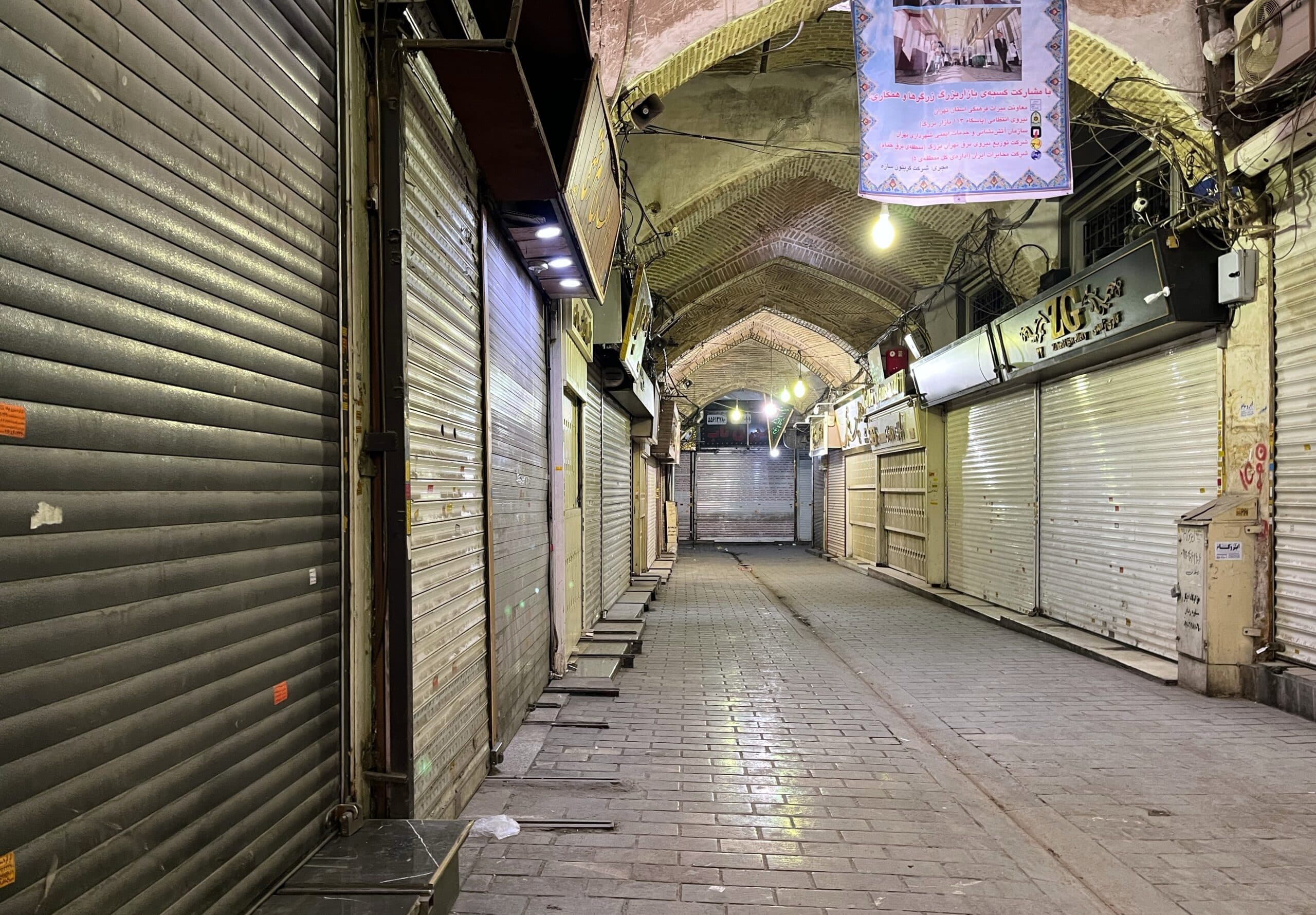 A quiet, covered bazaar corridor with closed metal shutters lining both sides and a few lit hanging lamps overhead.