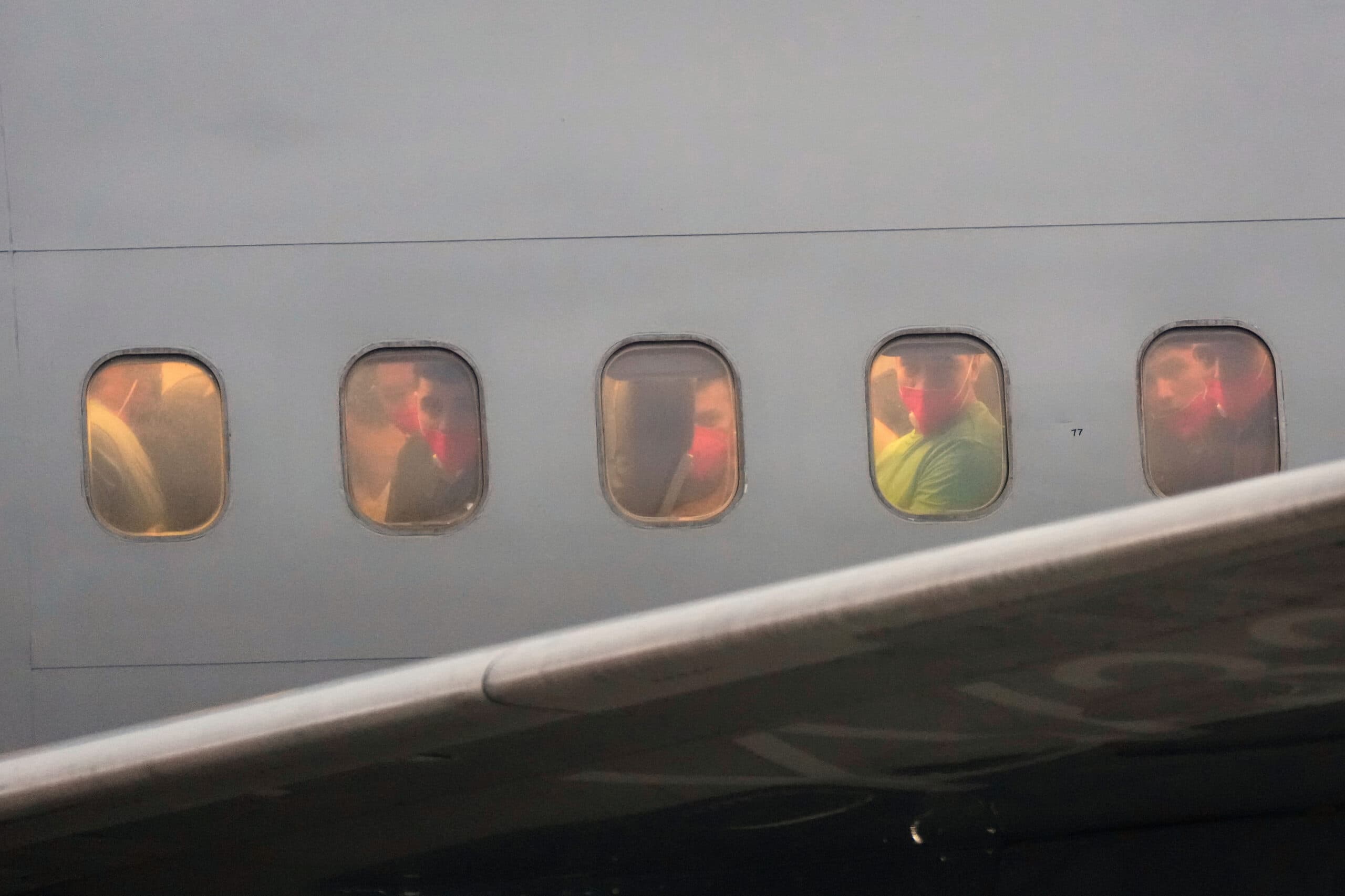 Passengers wearing face masks are visible through the windows of an airplane, with a wing partially covering the view.