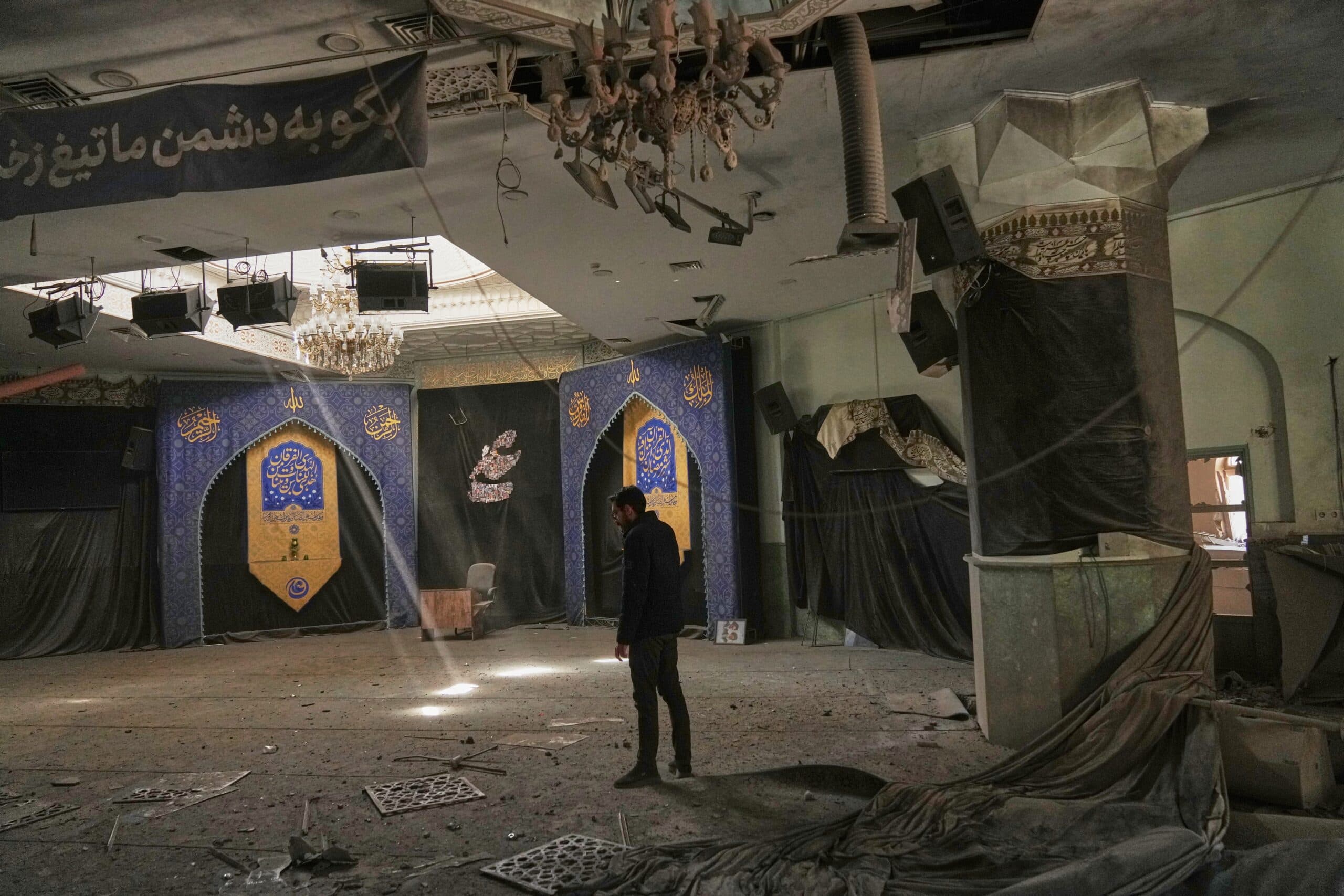 A man stands in a damaged room with debris on the floor, broken ceiling panels, and torn drapes. The walls feature ornate blue and gold arches with Arabic script, indicating a religious setting.