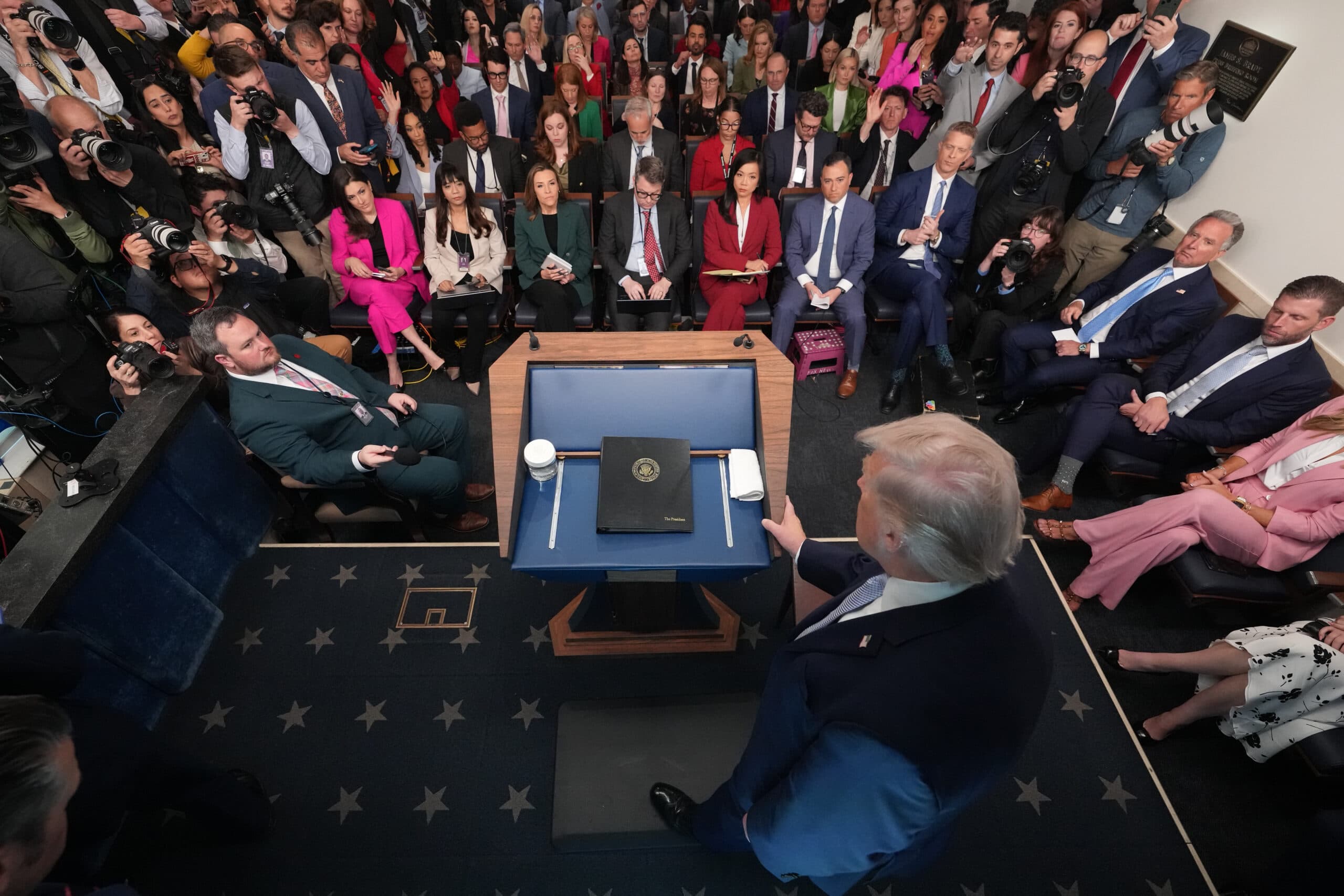 A man stands at a podium in a packed room with seated and standing attendees, many holding cameras.