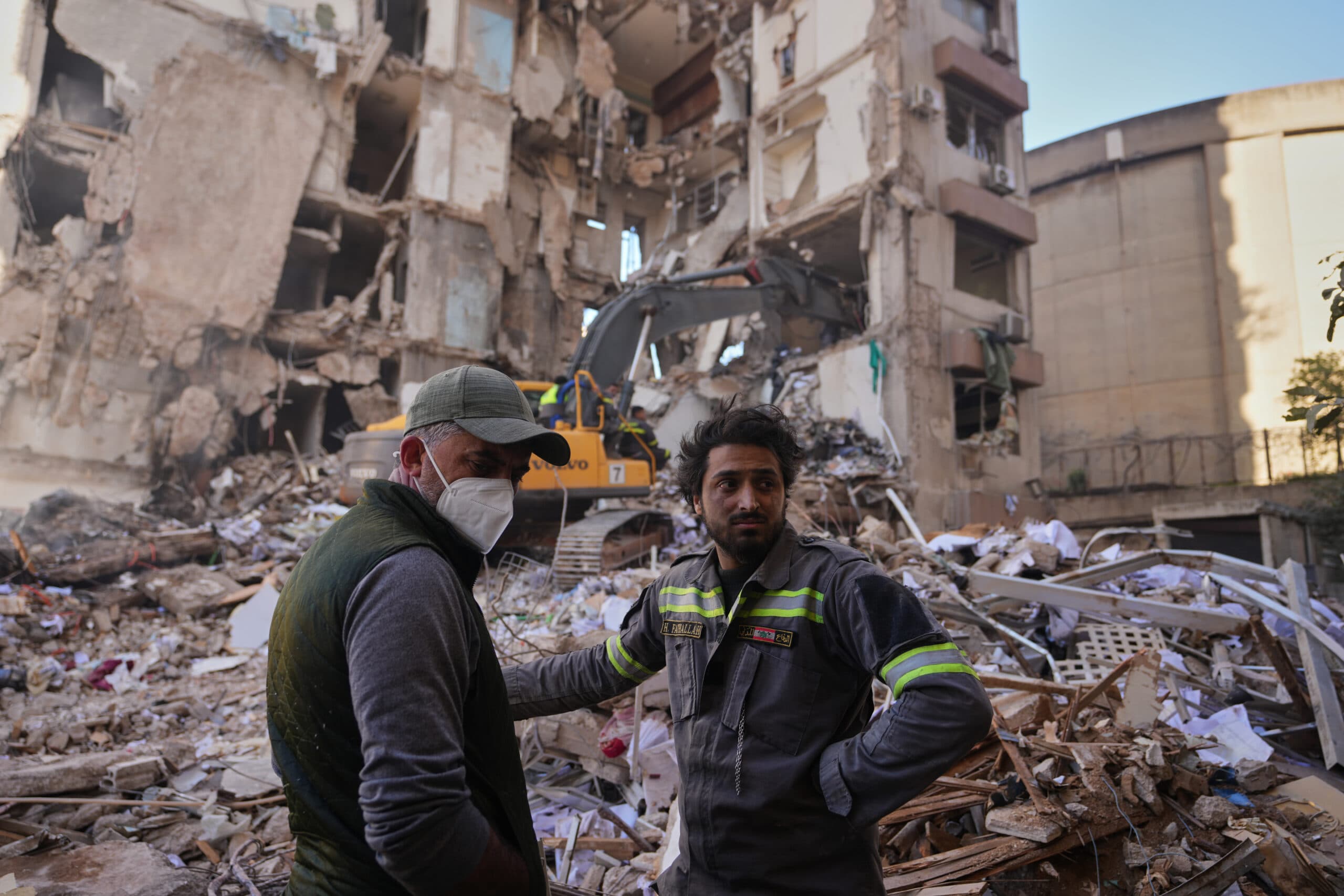 Two men stand amidst rubble and debris in front of a partially collapsed building, with an excavator in the background.