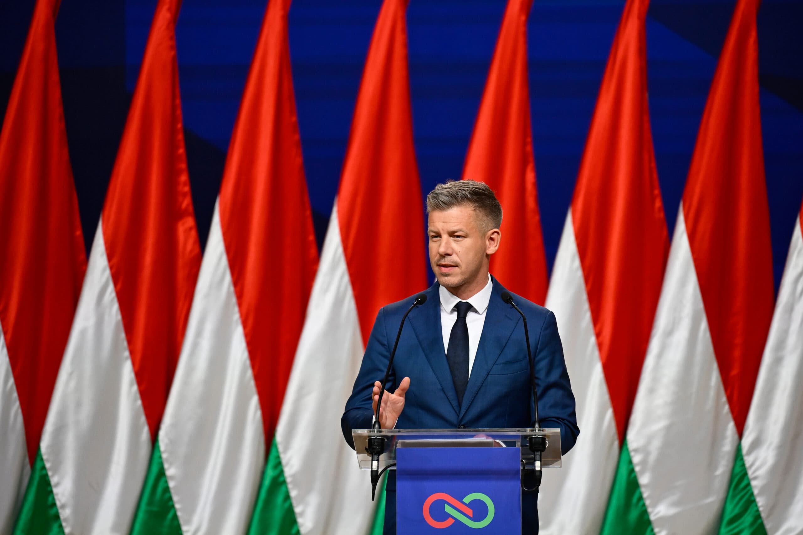 A man in a blue suit speaks at a podium with multiple red, white, and green flags in the background.