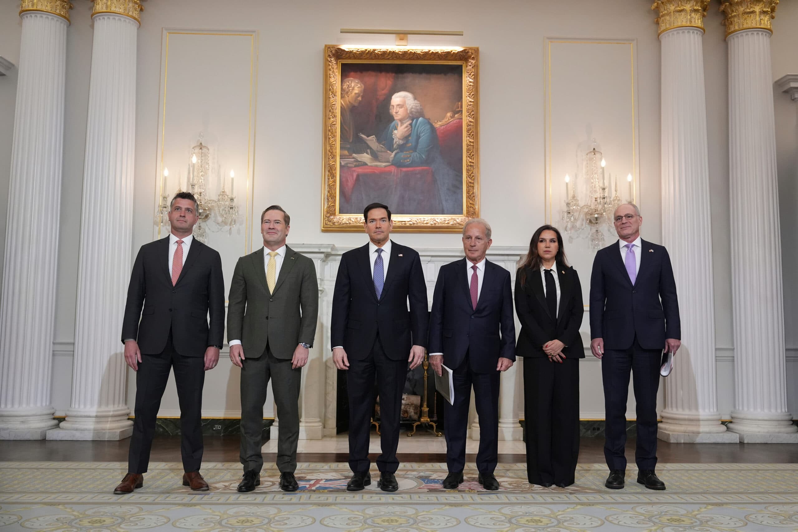 Six individuals stand in formal attire inside an elegant room with a large painting and ornate decor.