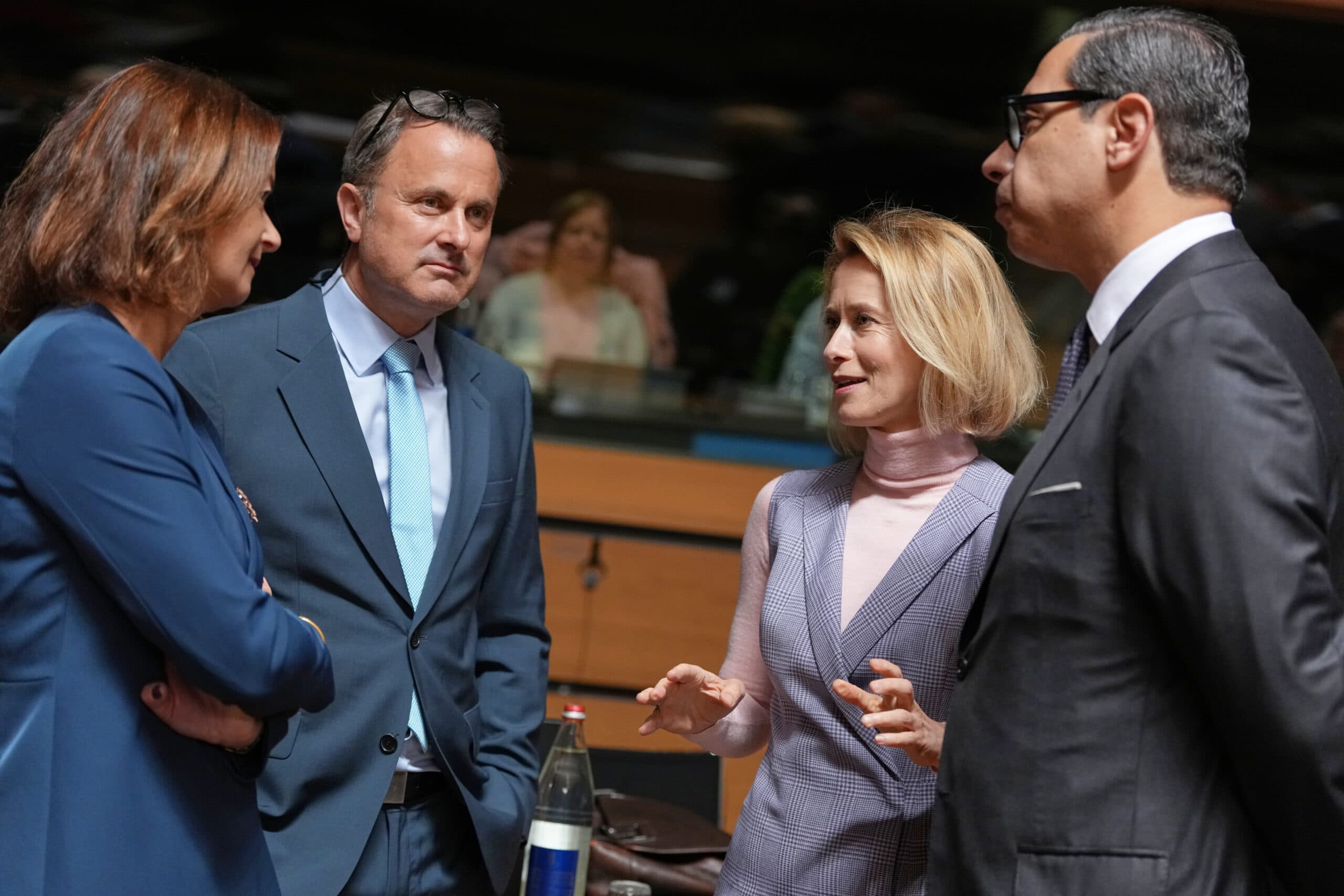 Four people in formal attire engaged in a discussion indoors, with a blurred audience in the background.