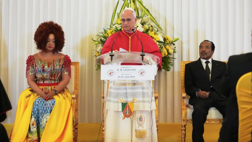 A cardinal in red robes speaks at a podium flanked by a woman in a colorful dress and a man in a black suit.
