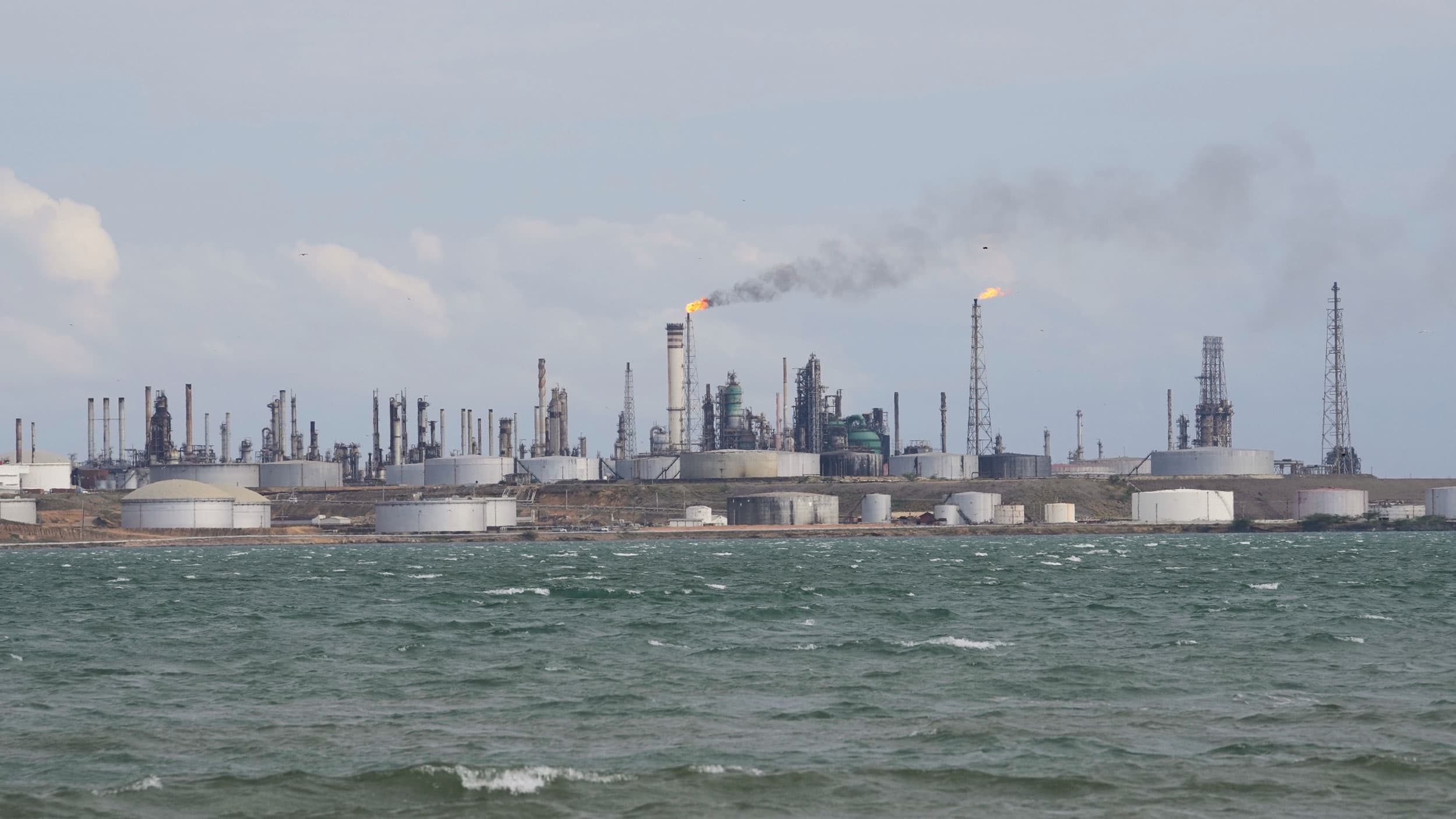 An industrial refinery complex by the sea with tall smokestacks emitting smoke and large storage tanks in the foreground.