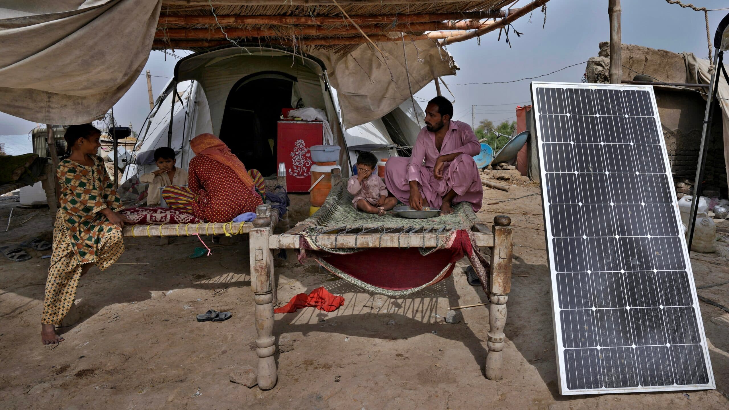 A family sits on woven cots under a thatched shelter with a solar panel nearby, in what appears to be a makeshift outdoor setting.