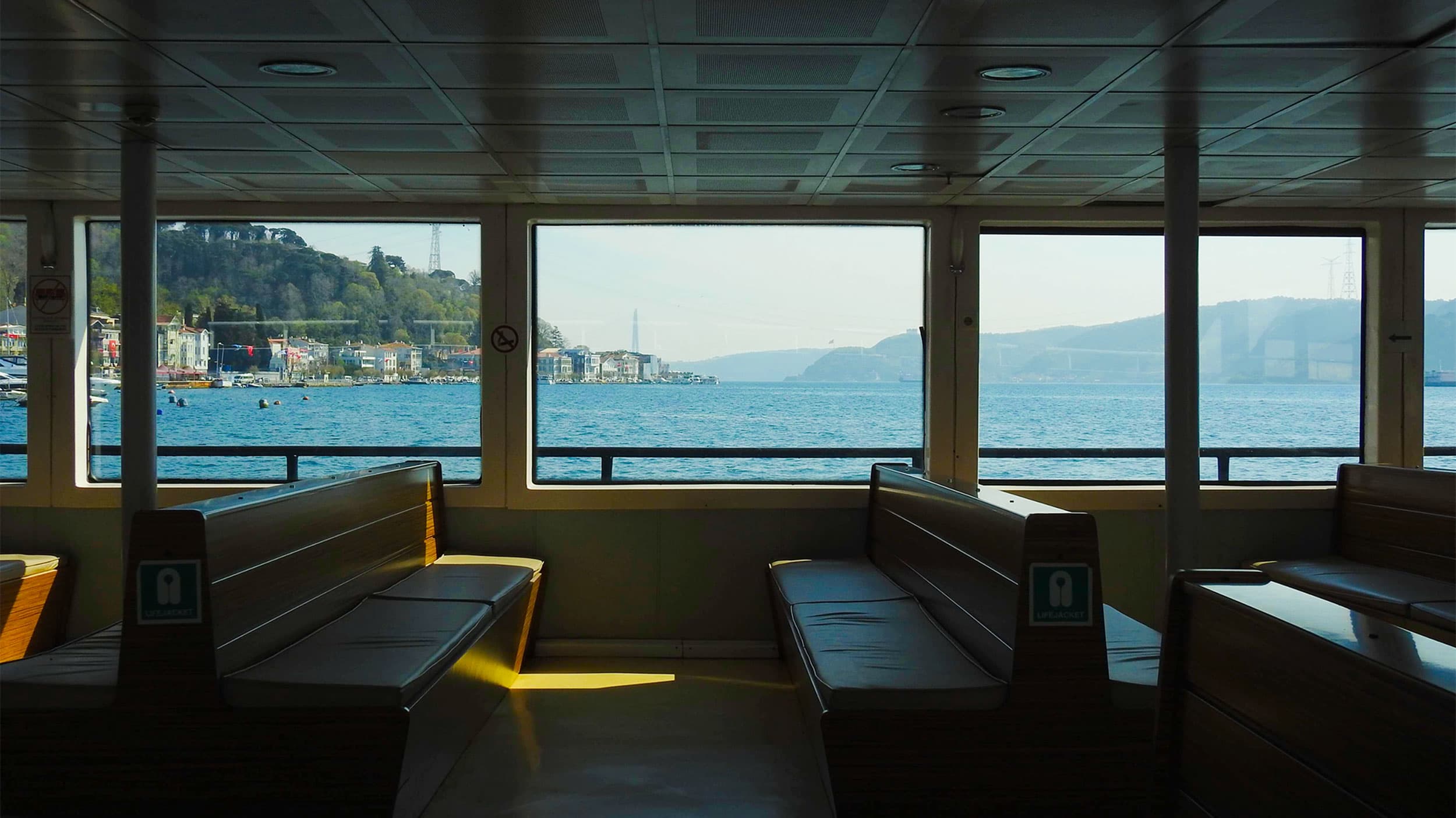 Inside a ferry, two rows of benches face large windows offering a view of a calm sea and distant coastline.