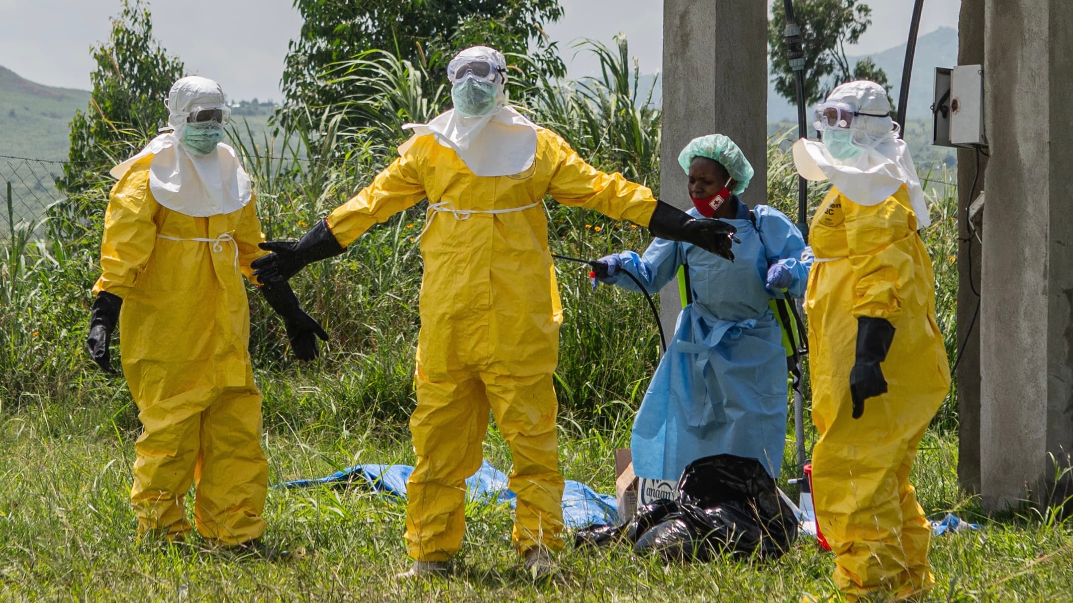 Four people in yellow hazmat suits and masks stand on grass, with one person in blue protective gear assisting.