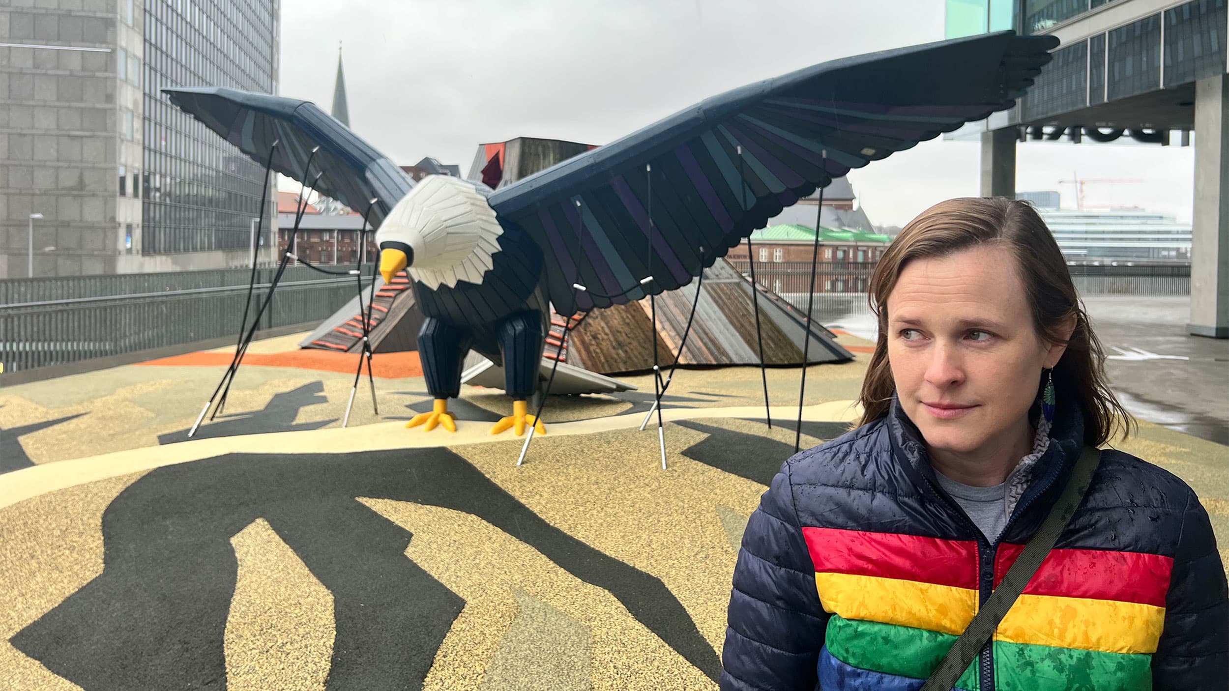 A person stands in front of a large sculpture of an eagle with outstretched wings on a rooftop.