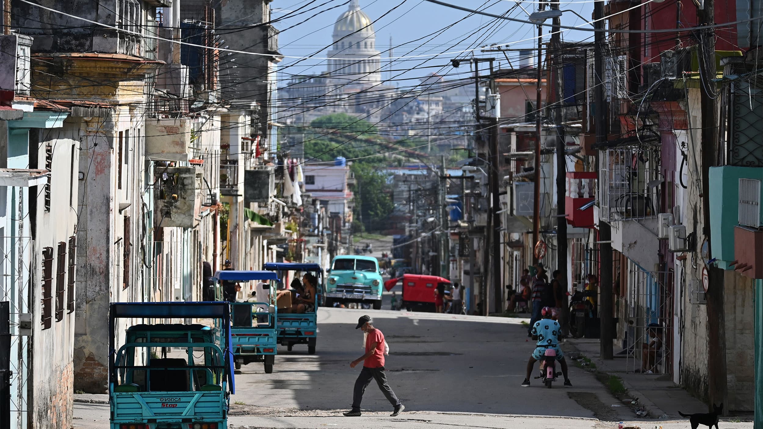 A street scene with people walking, auto rickshaws, and buildings. A dome structure is visible in the background.