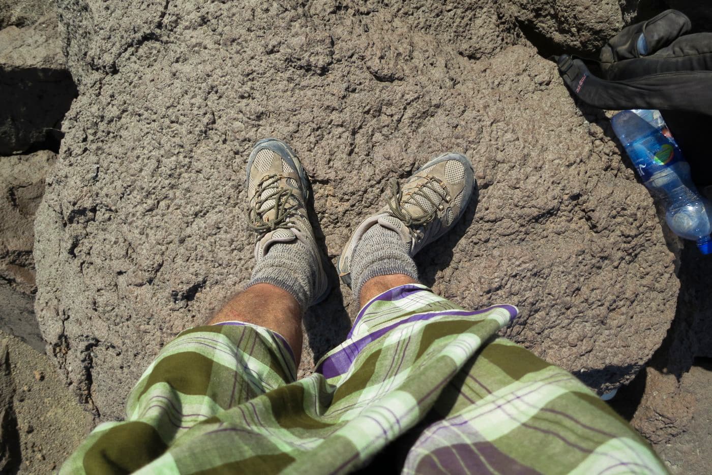 Person wearing hiking boots and plaid pants standing on rocky terrain with a water bottle and backpack nearby.