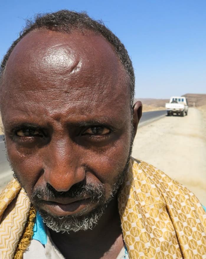 A man with short hair and a beard stands near a road with a truck in the distance on a sunny day.
