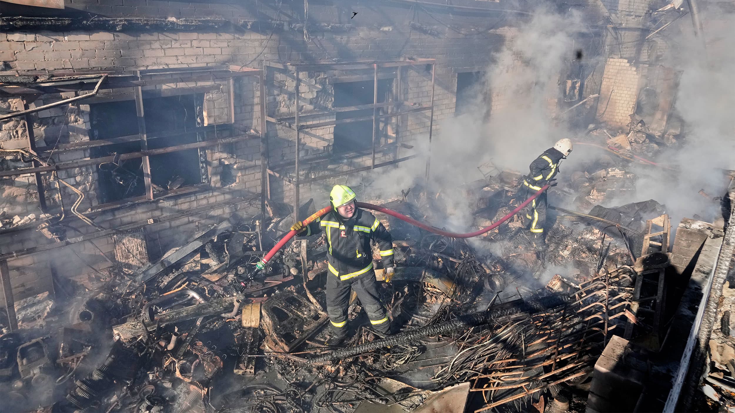 Two firefighters in protective gear work amidst smoldering debris in a heavily damaged building.
