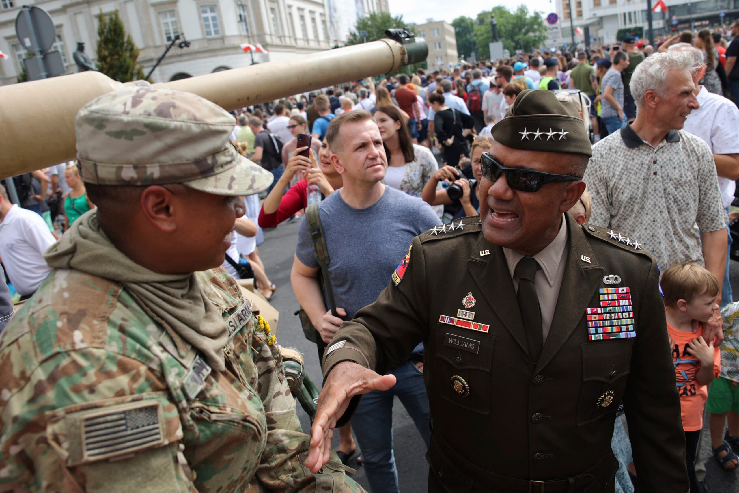 Two military personnel in uniform greet each other amidst a crowd, with onlookers taking photos.