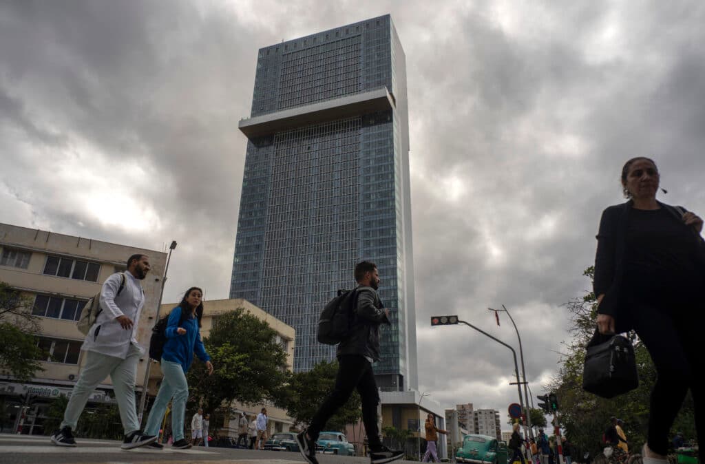 People walking near a tall, modern skyscraper under a cloudy sky.