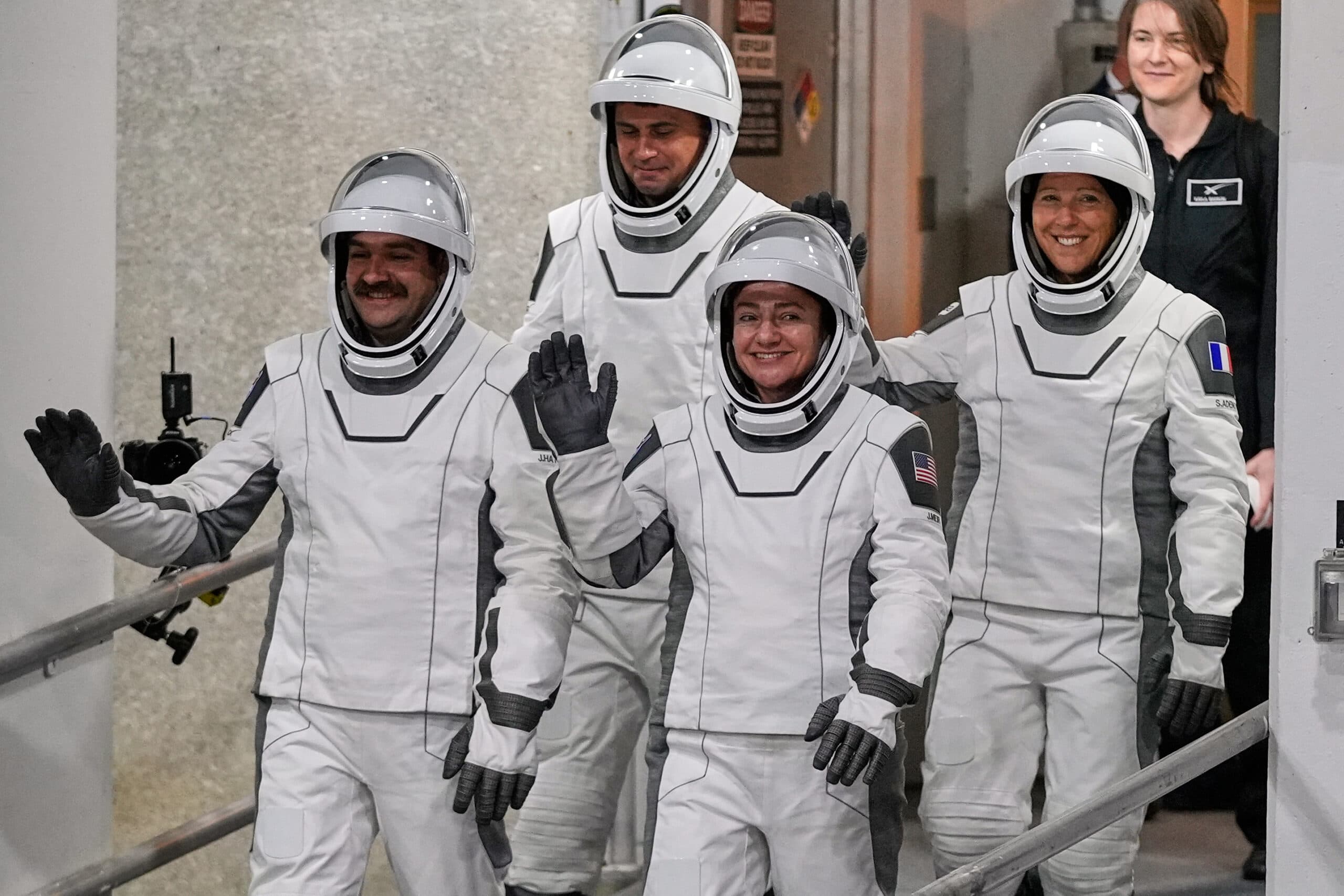 Four astronauts in white space suits with helmets, waving and walking down a hallway, with a person observing in the background.
