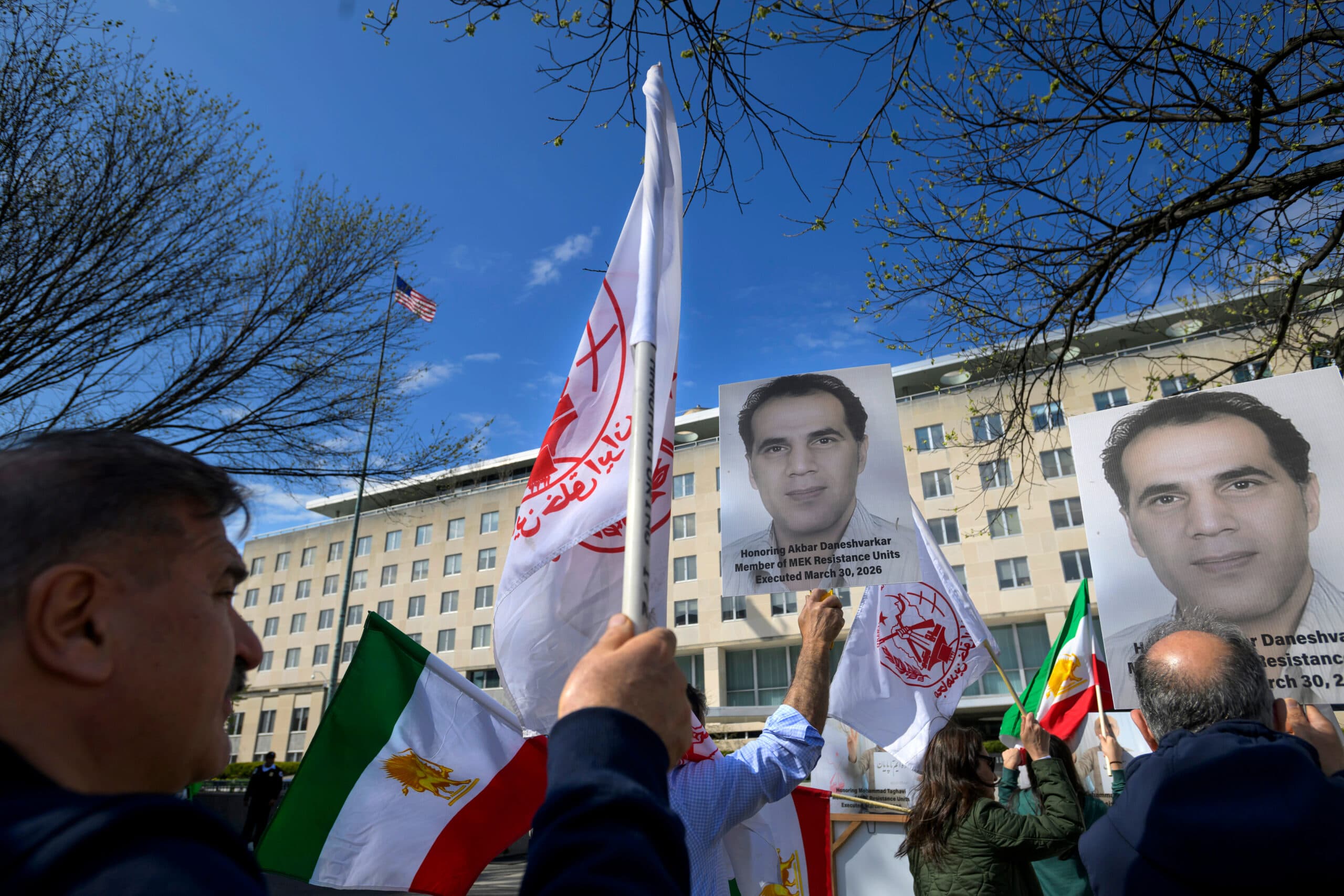 People holding flags and portraits in a protest outside a large government building, with trees and a clear sky in the background.