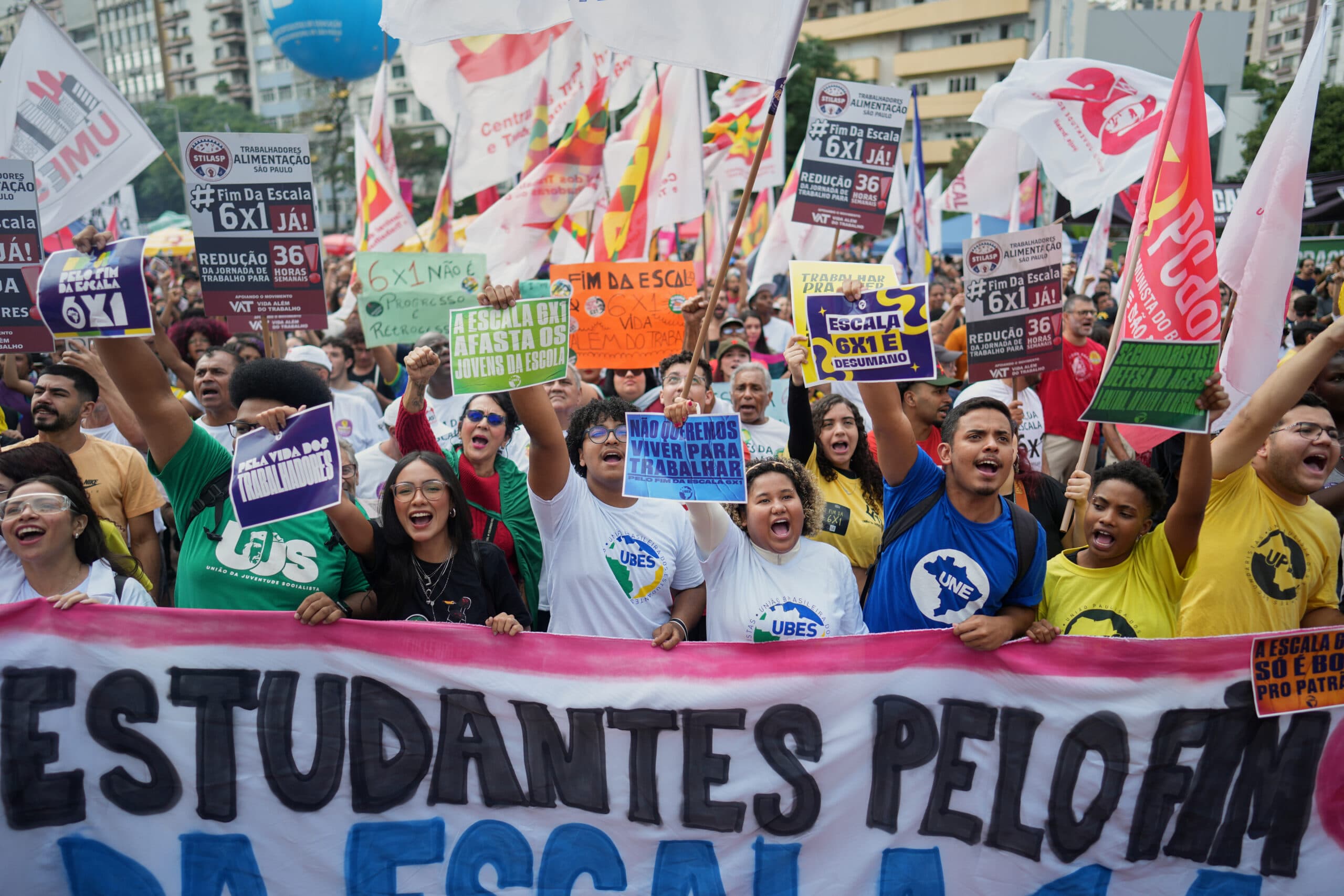 A group of people are gathered at a protest, holding colorful signs and banners in a city setting.