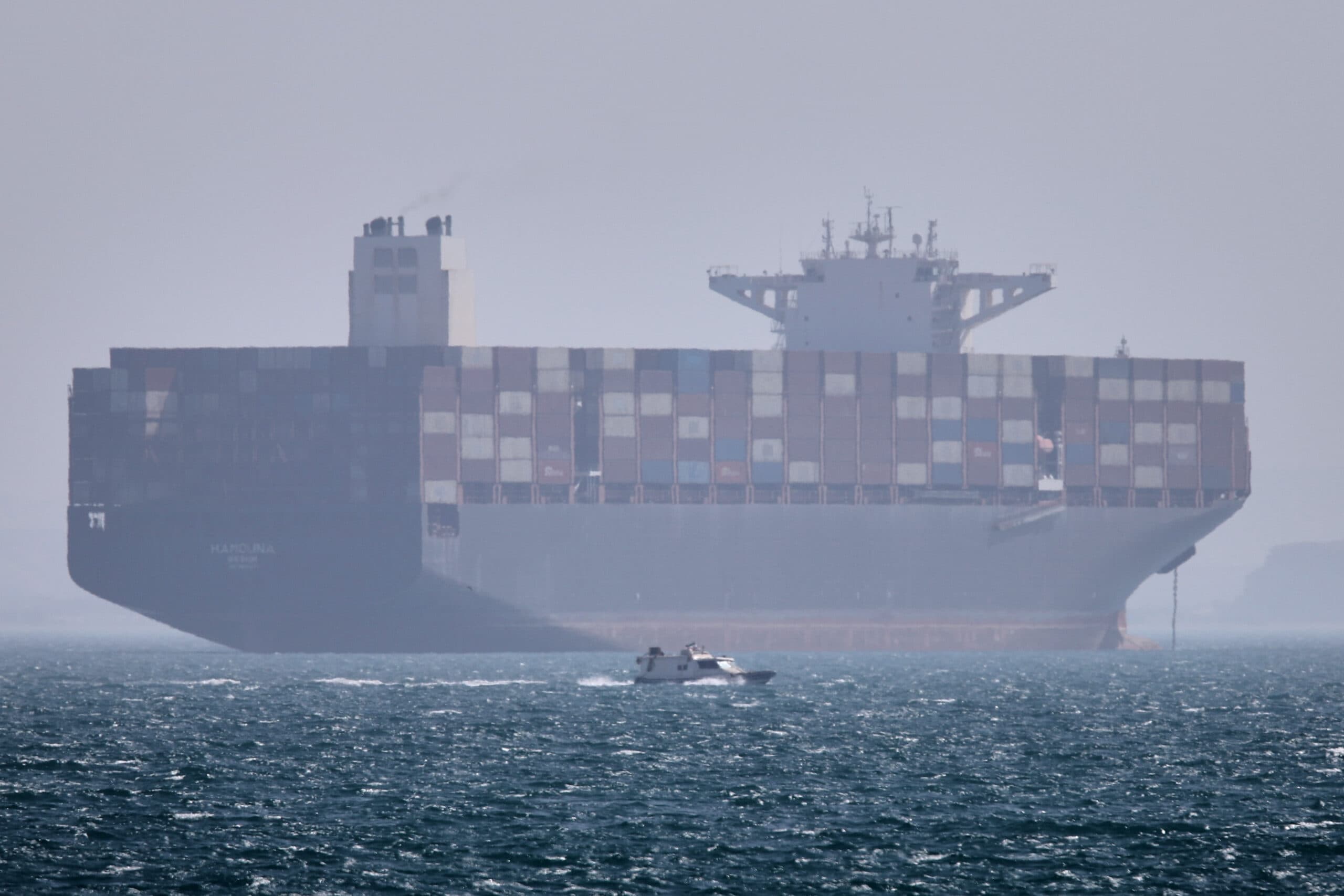 A large container ship loaded with colorful cargo containers is sailing on a foggy sea, with a small boat in the foreground.