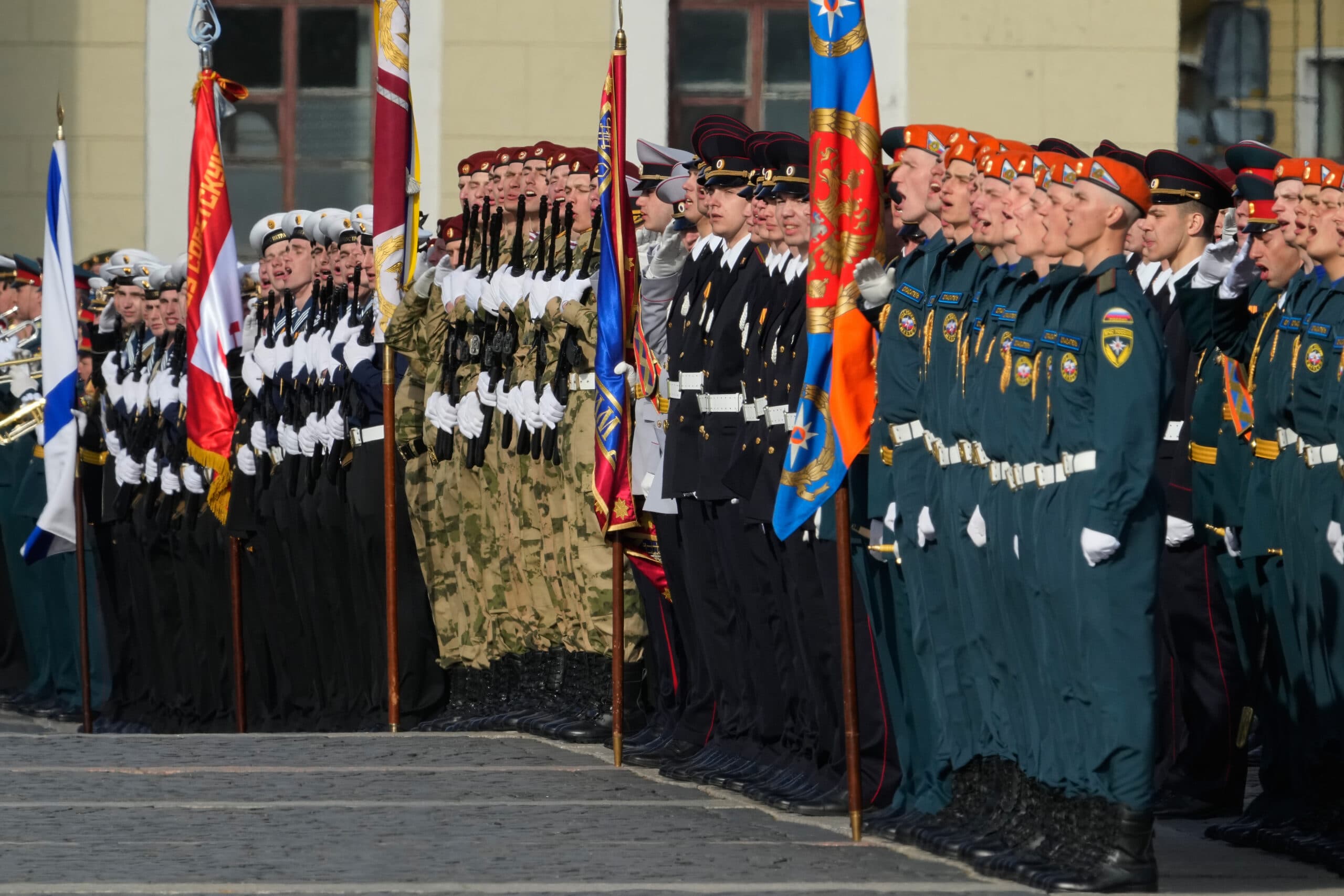 A line of uniformed soldiers stands at attention, each holding colorful flags, in a formal ceremony setting.