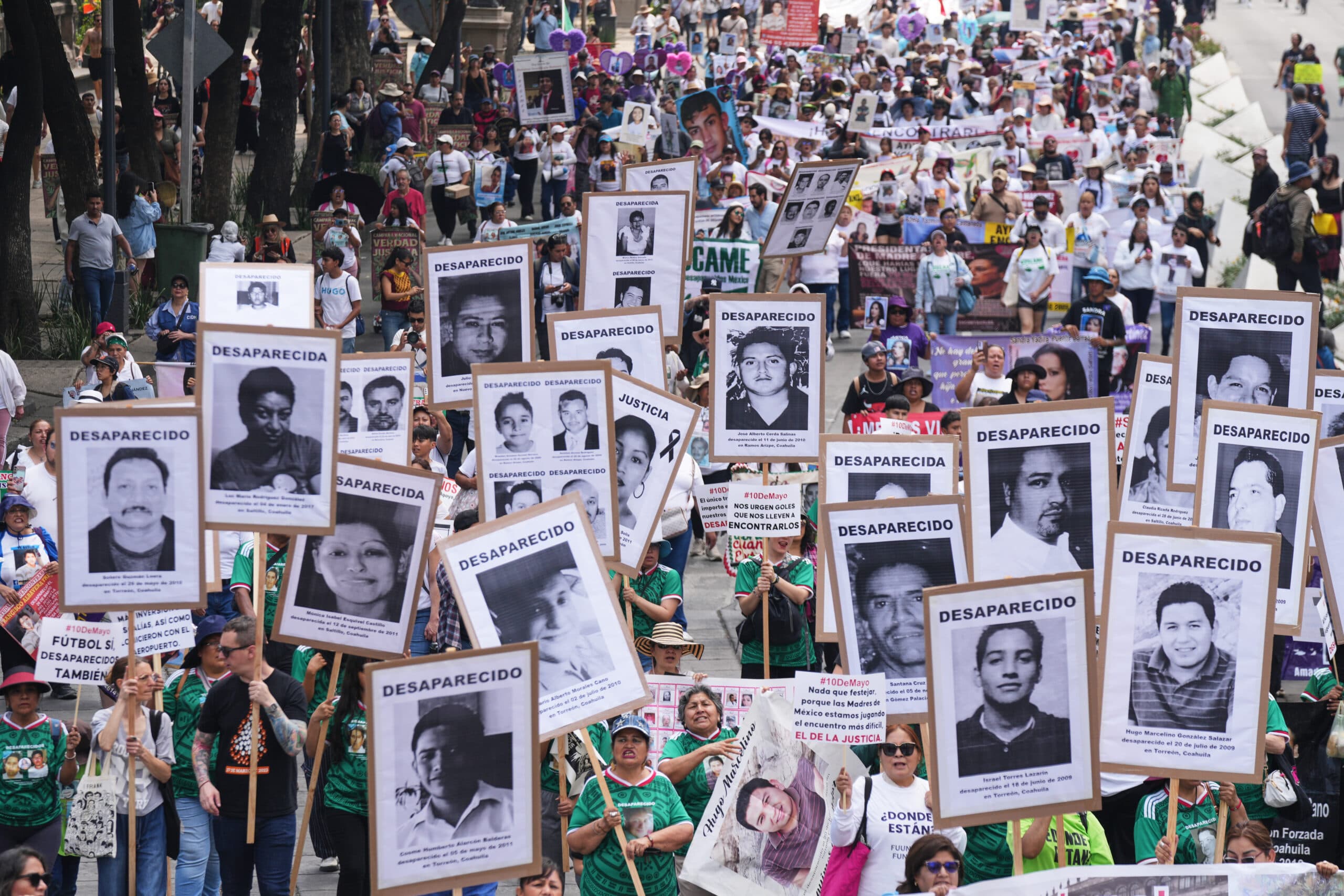 A large crowd holds up signs with photos of missing persons, marked "DESAPARECIDO," during a protest or march.