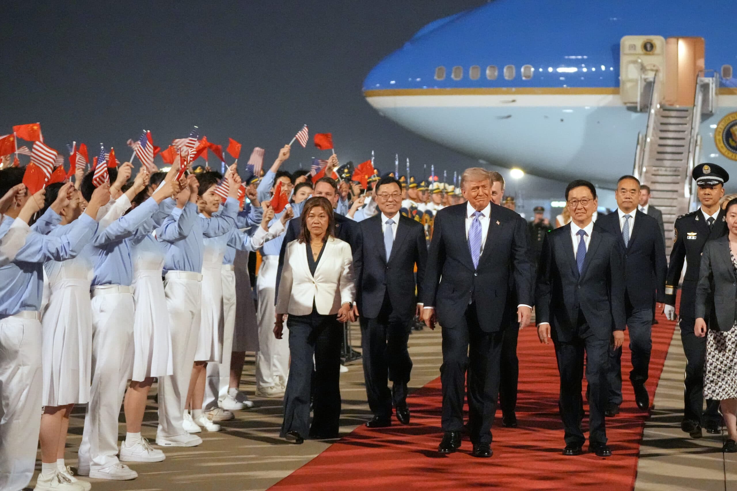 A group of dignitaries walking on a red carpet with an aircraft in the background, while a crowd waves flags.