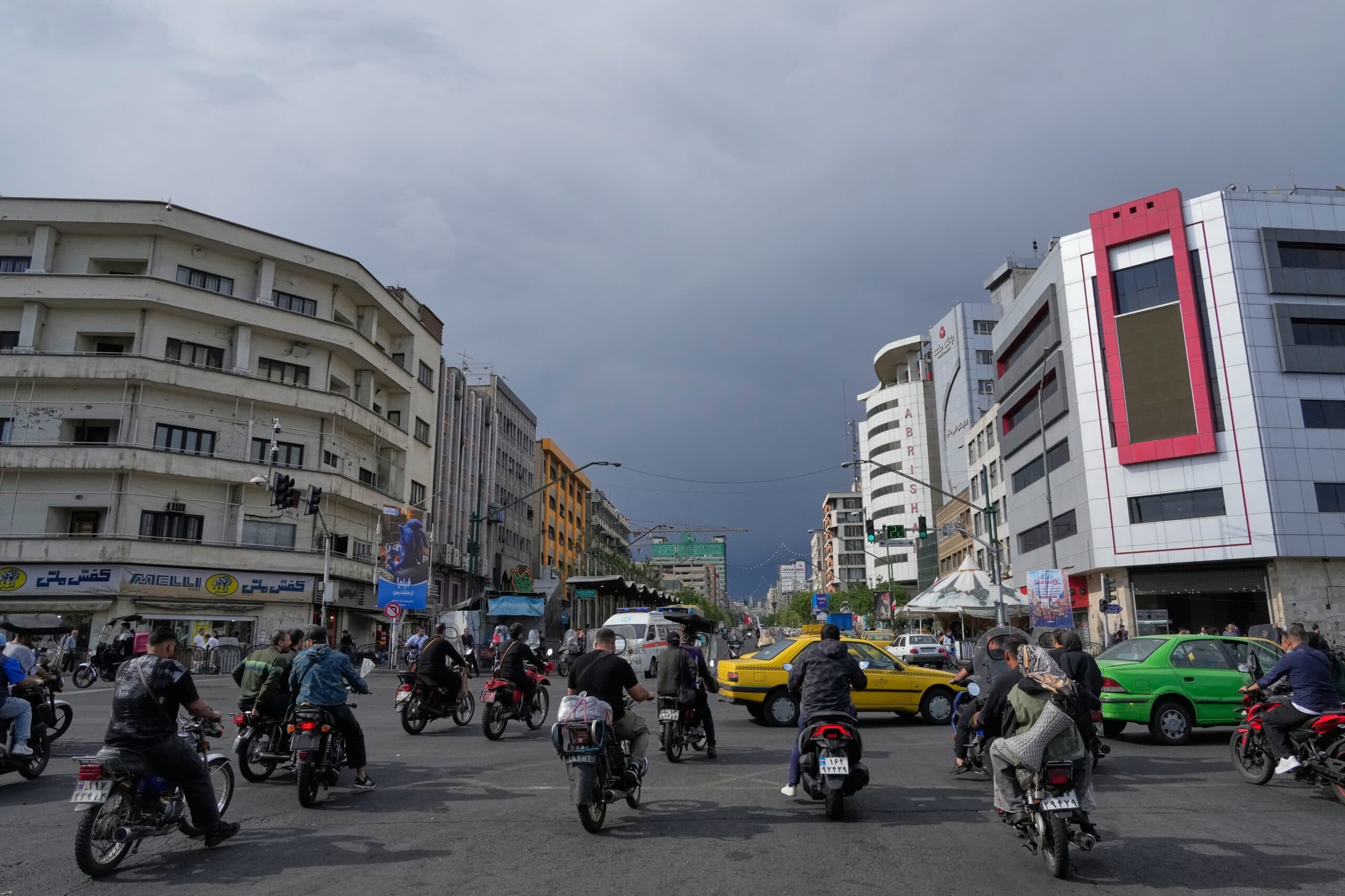 A busy urban street with numerous motorcyclists and cars under a cloudy sky, surrounded by multi-story buildings.